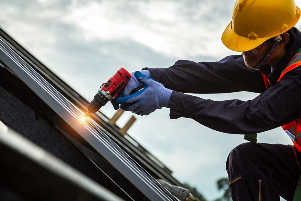 Two workers install light-colored shingles on a sloped roof under sunny skies.