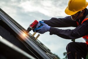 Two workers install light-colored shingles on a sloped roof under sunny skies.