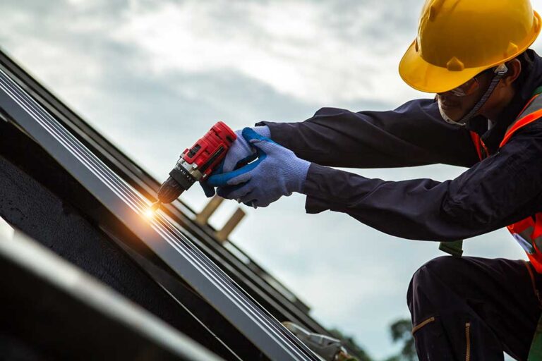 Two workers install light-colored shingles on a sloped roof under sunny skies.